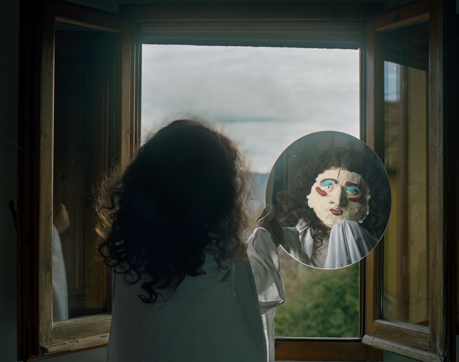 A figure at an open window holds a round mirror that shows a painted mask-like face