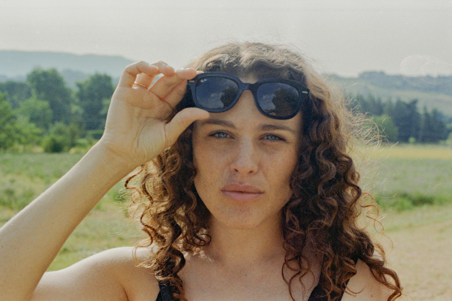 Portrait of Claire Pirovano lifting her sunglasses, curly hair, in a green landscape