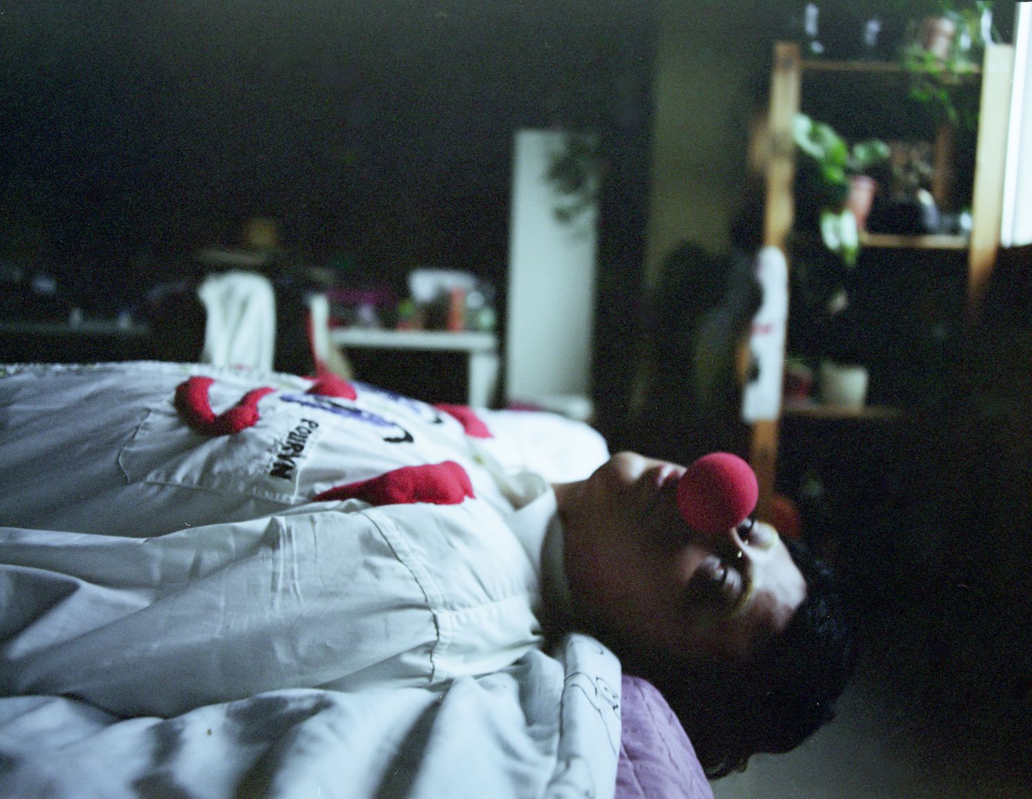 A person lying on a bed wearing a red clown nose, seen sideways